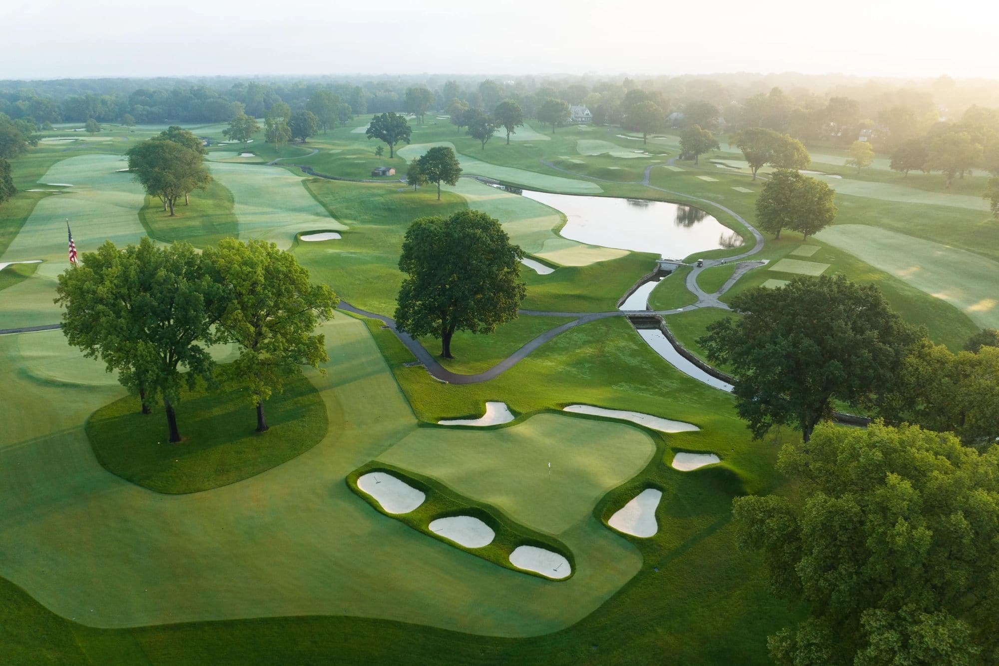 Aerial view of a manicured golf green with bunkers, a pond, and tree-lined fairways in golden morning light